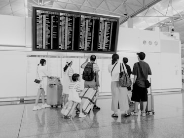A group of people stand in front of a large flight information board at an airport. They are looking at the departure and arrival details. The scene is monochrome, and several individuals have luggage with them. The architecture of the space includes high ceilings and industrial elements.