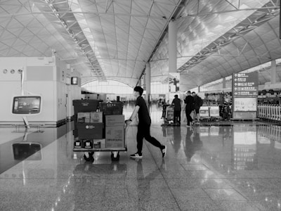 An airport terminal with a high, geometric ceiling design and reflective floors. A person is pushing a cart filled with boxes and packages, while other individuals are gathered in the background near check-in counters. There are signs in different languages and luggage trolleys lined up to the right.