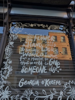 A storefront window displays a quote by Georgia O'Keeffe, written in white script and surrounded by floral designs. The writing is clear against a dark background, with reflections of a brick building visible in the glass.