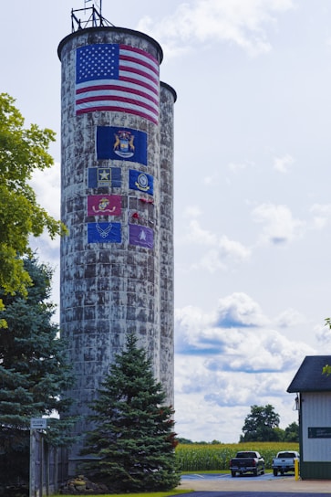 A tall, weathered silo displays an American flag at the top, with several military insignia and logos painted below it. Surrounding the silo are lush green trees and an open field. Two vehicles are parked nearby, next to a white building. The sky is partly cloudy, creating a serene and patriotic scene.