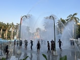 A lively scene in a water park with people enjoying water fountains spraying from overhead structures. The area is surrounded by tall palm trees, and the sky is clear with a light blue hue.