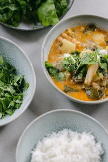 A close-up shot of a meal consisting of different dishes in bowls. One bowl contains a colorful stew with vegetables, herbs, and sesame seeds. Surrounding it, there are bowls with fresh leafy greens and chopped herbs, and a bowl of plain white rice.