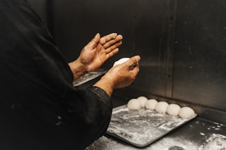 A person wearing a dark outfit is shaping dough with their hands over a metallic countertop. Several dough balls are neatly arranged on a floured baking tray beside them.