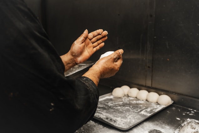 A person wearing a dark outfit is shaping dough with their hands over a metallic countertop. Several dough balls are neatly arranged on a floured baking tray beside them.