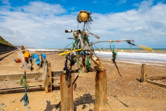 A sculpture made from driftwood, plastic, and other debris stands on a sandy beach. The structure is mounted between two posts and resembles a humanoid figure. In the background, the sea meets the sandy shore under a partly cloudy sky. Various colorful plastic items are used to construct the figure, including bottles, bags, and ropes.