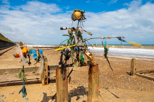 A sculpture made from driftwood, plastic, and other debris stands on a sandy beach. The structure is mounted between two posts and resembles a humanoid figure. In the background, the sea meets the sandy shore under a partly cloudy sky. Various colorful plastic items are used to construct the figure, including bottles, bags, and ropes.