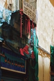 A rustic street scene featuring a small balcony with a wire mesh and a hand-painted sign displaying art services. Bright red high-heeled boots hang from the railing, contrasted by blue netting draped across the facade. The building has a rough, textured wall and a shop sign visible below.