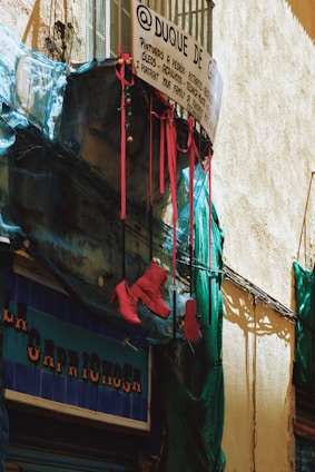 A rustic street scene featuring a small balcony with a wire mesh and a hand-painted sign displaying art services. Bright red high-heeled boots hang from the railing, contrasted by blue netting draped across the facade. The building has a rough, textured wall and a shop sign visible below.