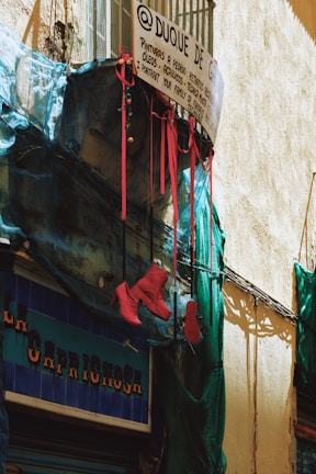 A rustic street scene featuring a small balcony with a wire mesh and a hand-painted sign displaying art services. Bright red high-heeled boots hang from the railing, contrasted by blue netting draped across the facade. The building has a rough, textured wall and a shop sign visible below.