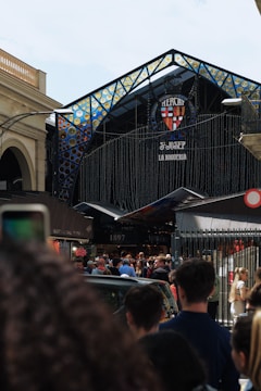 A bustling scene at the entrance of a market, with a large crowd gathered beneath a decorative, stained-glass-style archway. The market's entrance is adorned with intricate designs, and a sign displays 'Mercat St. Josep La Boqueria'. The crowd includes people of various ages, dressed in casual attire, and the atmosphere seems lively and busy.