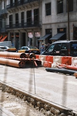 A city street scene with construction work in progress, featuring stacked orange and red pipes, and barriers. Several cars, including a taxi, are parked along the side of the road. A group of people is gathered near the storefronts in the background, which include signs for a supermarket.
