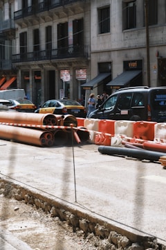 A city street scene with construction work in progress, featuring stacked orange and red pipes, and barriers. Several cars, including a taxi, are parked along the side of the road. A group of people is gathered near the storefronts in the background, which include signs for a supermarket.