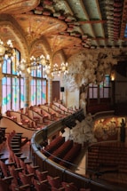 An ornate theater interior features a vibrant, intricately designed ceiling with stained glass windows and decorative arches. Rows of plush red seats curve elegantly around the theater, accompanied by artistic sculptures and elaborate light fixtures hanging from above.