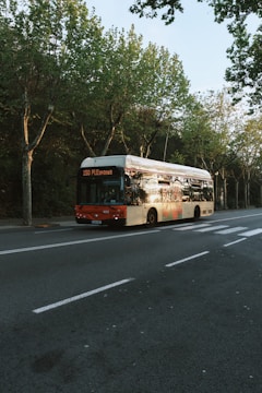 A bus with the number 150 and destination PLEspanya is parked on a tree-lined street. The bus has a red front and an advertisement on its side. The roadway is empty, with visible lanes and a bicycle lane on the side. Leafy green trees line the sidewalk, casting shadows on the street.