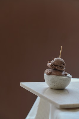 A small, speckled gray bowl holds two scoops of chocolate ice cream, with a stick inserted in the top. The bowl is placed on a cream-colored, triangular stone surface against a plain brown background.
