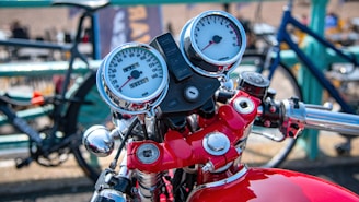 Chrome motorcycle handlebars with dual round gauges displaying speed and RPM. The bike's frame is a vibrant red, and a background of blurred bicycles with a brightly colored fence adds depth.
