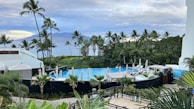 A tropical resort scene with a round swimming pool surrounded by lounge chairs and umbrellas, set amidst lush greenery and palm trees. In the background, there is a view of the ocean and a distant mountainous landscape under a partly cloudy sky.