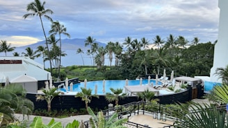 A tropical resort scene with a round swimming pool surrounded by lounge chairs and umbrellas, set amidst lush greenery and palm trees. In the background, there is a view of the ocean and a distant mountainous landscape under a partly cloudy sky.