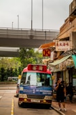 A red and blue public minibus with the destination Tsuen Wan is parked on the side of an urban street. A man is walking on the sidewalk nearby, talking on his phone. Above the street is a flyover or bridge. Several storefronts with signs written in Chinese characters, including a convenience store sign with a 'K', are visible on the right. In the background, there is greenery.