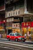 A bustling urban street scene with a bright red taxi driving past traditional shops with Chinese signage. People are standing on the sidewalk near a bus stop, and various storefronts display goods inside their glass windows. The multi-story buildings have an aged appearance, adding to the city's rich cultural atmosphere.