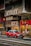 A bustling urban street scene with a bright red taxi driving past traditional shops with Chinese signage. People are standing on the sidewalk near a bus stop, and various storefronts display goods inside their glass windows. The multi-story buildings have an aged appearance, adding to the city's rich cultural atmosphere.