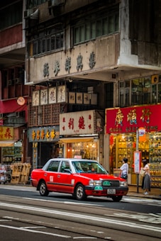 A bustling urban street scene with a bright red taxi driving past traditional shops with Chinese signage. People are standing on the sidewalk near a bus stop, and various storefronts display goods inside their glass windows. The multi-story buildings have an aged appearance, adding to the city's rich cultural atmosphere.