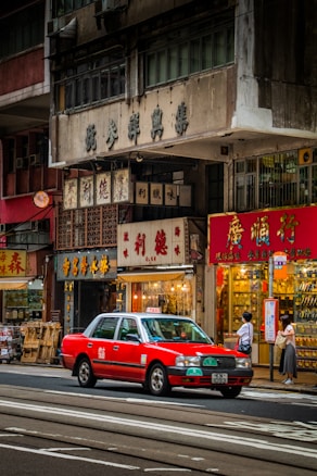 A bustling urban street scene with a bright red taxi driving past traditional shops with Chinese signage. People are standing on the sidewalk near a bus stop, and various storefronts display goods inside their glass windows. The multi-story buildings have an aged appearance, adding to the city's rich cultural atmosphere.