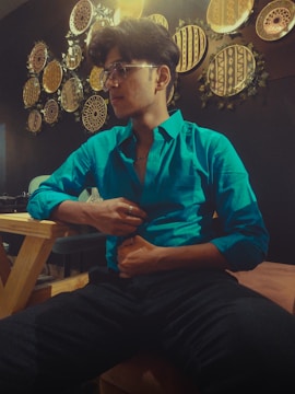 A person in a bright turquoise shirt sits on a wooden chair in a warmly lit room. The wall behind them is decorated with an array of circular, tribal-patterned art pieces with earthy tones. They are wearing glasses and seem relaxed, with one hand touching their shirt.