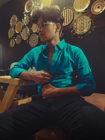 A person in a bright turquoise shirt sits on a wooden chair in a warmly lit room. The wall behind them is decorated with an array of circular, tribal-patterned art pieces with earthy tones. They are wearing glasses and seem relaxed, with one hand touching their shirt.