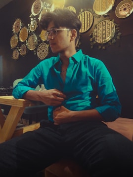 A person in a bright turquoise shirt sits on a wooden chair in a warmly lit room. The wall behind them is decorated with an array of circular, tribal-patterned art pieces with earthy tones. They are wearing glasses and seem relaxed, with one hand touching their shirt.