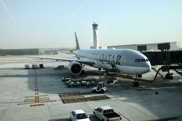 A large commercial airplane is stationed on an airport tarmac, connected to a passenger boarding bridge. Several ground crew members in high-visibility vests are gathered around service vehicles and equipment next to the aircraft. The airport control tower is visible in the background, under a clear sky.