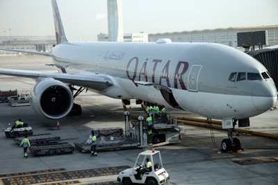 A large commercial airplane on the tarmac at an airport, with the word 'Qatar' prominently displayed on its fuselage. Ground crew members in high-visibility vests are busy with luggage handling and other aircraft preparations. The airport terminal and equipment are visible in the background.