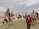 A group of people riding camels led by a guide in a desert landscape. The sky is overcast with clouds. The riders are wearing varied attire, some including scarves, hats, and sunglasses for protection against the sun and sand. The camels are fitted with colorful saddles and blankets.