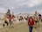 A group of people riding camels led by a guide in a desert landscape. The sky is overcast with clouds. The riders are wearing varied attire, some including scarves, hats, and sunglasses for protection against the sun and sand. The camels are fitted with colorful saddles and blankets.