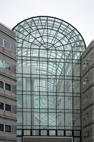 A modern architectural structure with a large glass atrium. The design features intricate geometric patterns made from black metal frames and transparent glass panels. Surrounding the atrium are stone-clad buildings with windows, creating a balance between glass and solid materials.