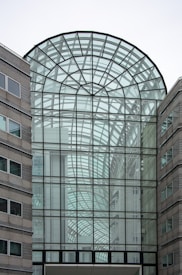 A modern architectural structure with a large glass atrium. The design features intricate geometric patterns made from black metal frames and transparent glass panels. Surrounding the atrium are stone-clad buildings with windows, creating a balance between glass and solid materials.