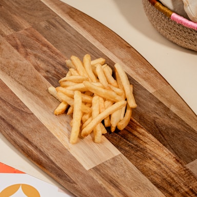 A pile of golden, crispy French fries sits on a wooden cutting board with varying shades of brown. The board's grain provides a rustic background. A woven basket with colorful accents is partially visible at the edge.