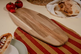 A wooden cutting board is placed on a striped red and mustard tablecloth surrounded by fresh tomatoes, a small red pepper, flatbreads, and a dish featuring a piece of flatbread with a topping. The scene conveys a rustic and cozy culinary setting.