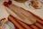 A wooden cutting board is placed on a striped red and mustard tablecloth surrounded by fresh tomatoes, a small red pepper, flatbreads, and a dish featuring a piece of flatbread with a topping. The scene conveys a rustic and cozy culinary setting.