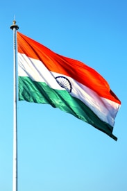 A large flag featuring three horizontal stripes of orange, white, and green, with a blue Ashoka Chakra wheel in the center. The flag is mounted on a tall flagpole against a clear blue sky.