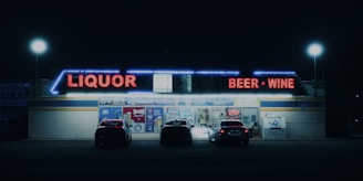 A nighttime scene of a liquor store with bright neon signs displaying 'Liquor' and 'Beer - Wine' in red lights. Several cars are parked in front of the store, which is illuminated by streetlights and its own lights. The store has a variety of advertisements and a neon 'Open' sign visible on the front.