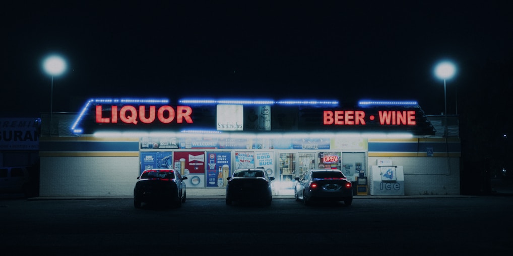 A nighttime scene of a liquor store with bright neon signs displaying 'Liquor' and 'Beer - Wine' in red lights. Several cars are parked in front of the store, which is illuminated by streetlights and its own lights. The store has a variety of advertisements and a neon 'Open' sign visible on the front.