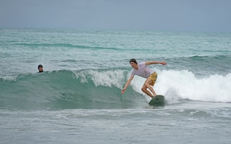 A surfer skillfully rides a wave on a light-colored surfboard while wearing a white t-shirt and yellow patterned shorts. Another person is visible in the water, watching from a distance with a helmet for protection. The scene is set on a calm sea with gentle waves under an overcast sky.