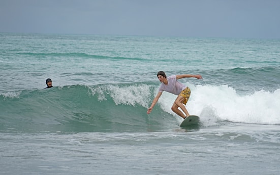 A surfer skillfully rides a wave on a light-colored surfboard while wearing a white t-shirt and yellow patterned shorts. Another person is visible in the water, watching from a distance with a helmet for protection. The scene is set on a calm sea with gentle waves under an overcast sky.