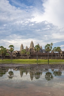 A historical temple complex is set against a backdrop of partially cloudy skies. The structure's intricate towers are surrounded by tall palm trees and reflect in a large body of water in the foreground.