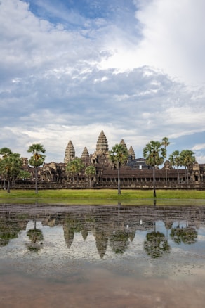A historical temple complex is set against a backdrop of partially cloudy skies. The structure's intricate towers are surrounded by tall palm trees and reflect in a large body of water in the foreground.