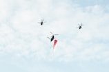 Three helicopters are flying in the sky carrying a large flag. The background is filled with light, fluffy clouds, suggesting a clear day.
