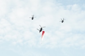 Three helicopters are flying in the sky carrying a large flag. The background is filled with light, fluffy clouds, suggesting a clear day.