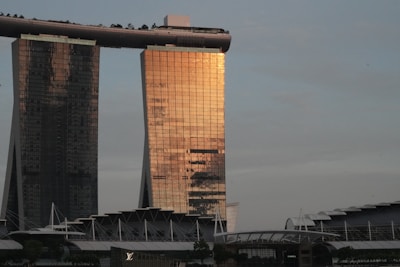 An iconic architectural structure with three tall towers connected by a large rooftop terrace or park. The building reflects a warm golden light from the sunset, highlighting its modern design. In the foreground, there is a complex of smaller buildings with a prominent designer store sign visible.