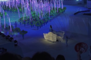 A vibrant performance scene with performers wearing costumes that resemble green leaves. The setting is illuminated with blue and purple lights, giving it a magical, ethereal feel. At the forefront, a person is dressed in a flowing gown adorned with lights, adding a touch of elegance to the scene.